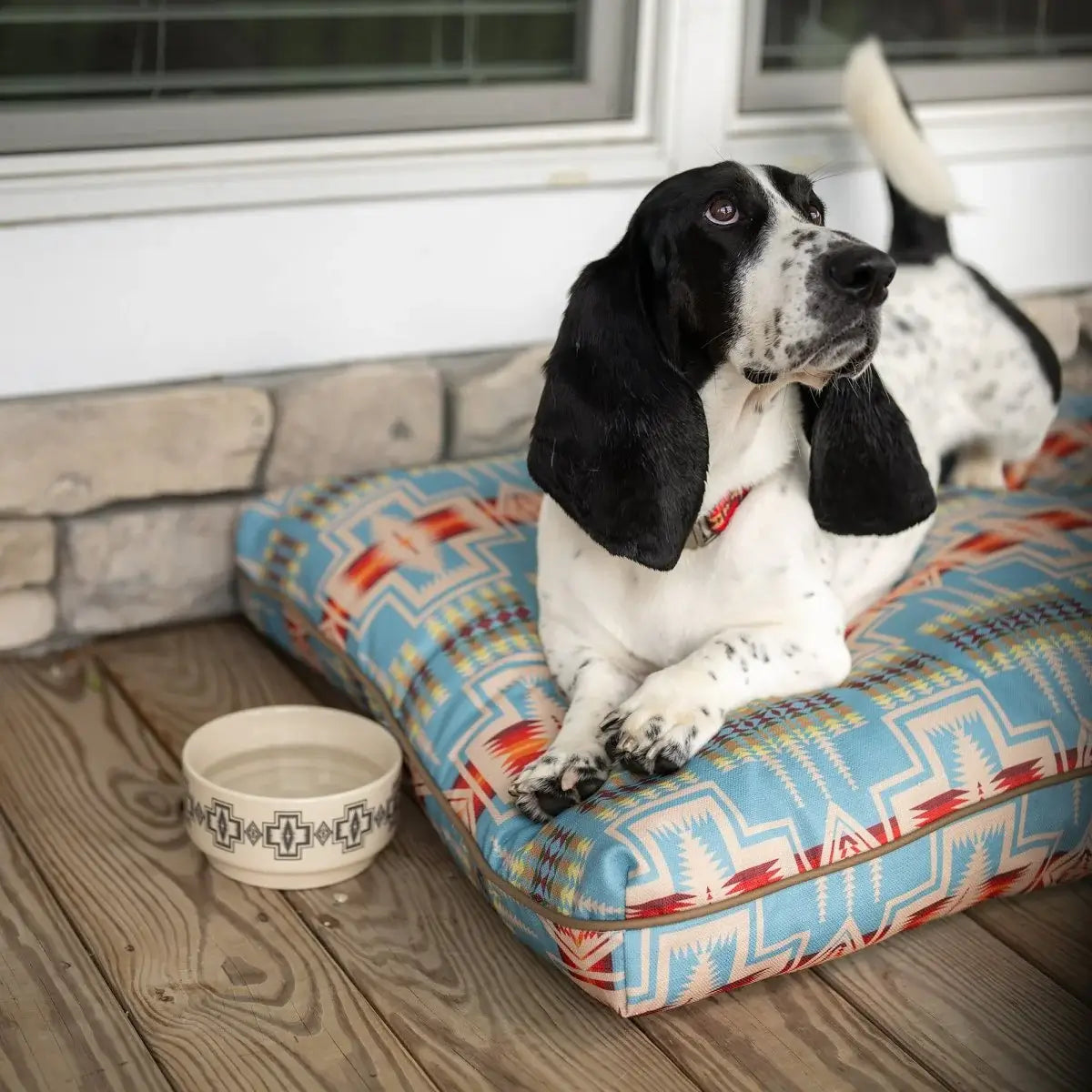 Black and white dog resting on colorful Pendleton dog bed with geometric pattern by a water bowl on wooden floor